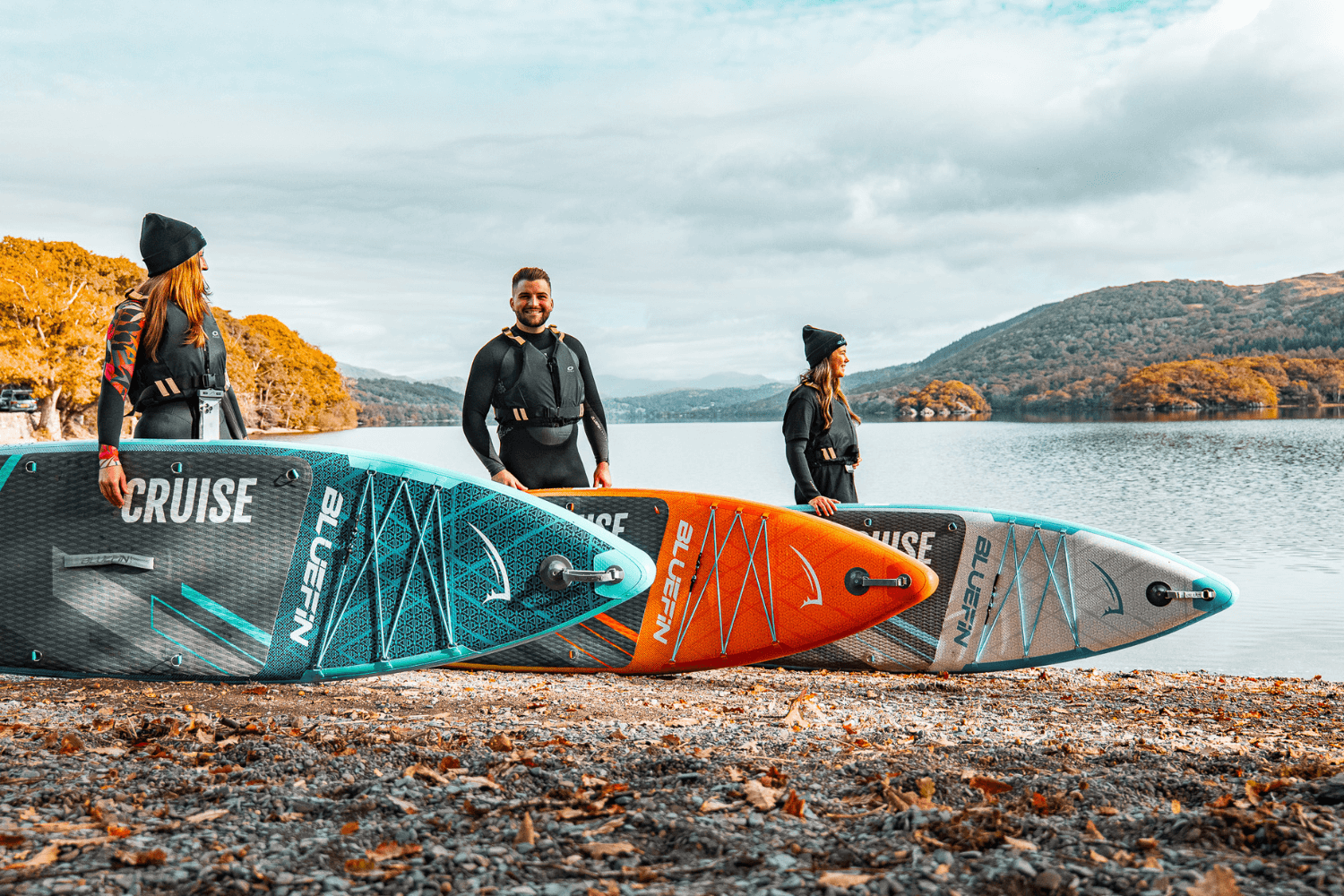 Three people standing by the water with inflatable paddleboards in blue, orange, and gray colors.