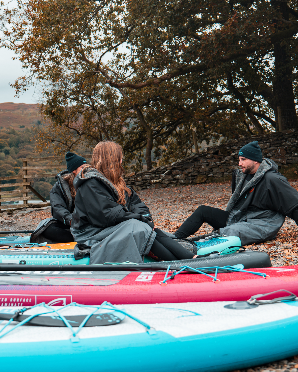 Group of people in black jackets sitting on colorful paddleboards by a rocky shore with autumn leaves.