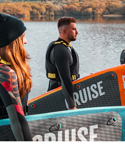 Two people in wetsuits holding inflatable paddleboards by a lake, autumn foliage in background