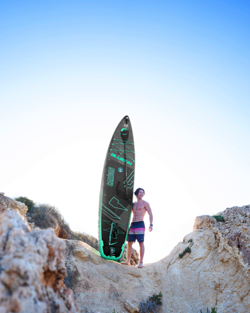 Man standing on rocks holding a black and green inflatable paddleboard against a clear blue sky