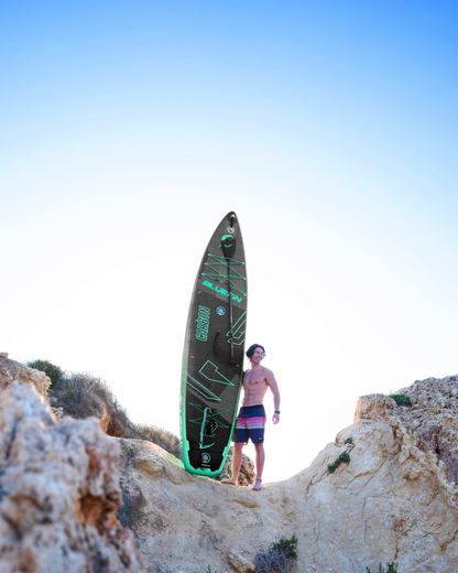 Man standing on rocks holding a black and green inflatable paddleboard against a clear blue sky