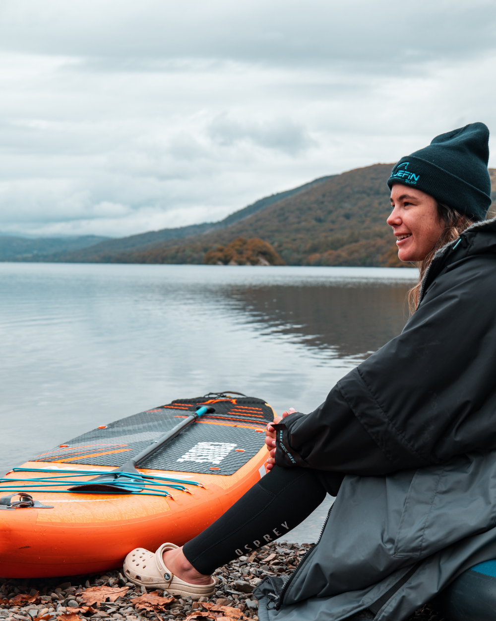 Woman sitting by a lake on gravel, wearing a beanie and rain jacket, with an orange paddleboard nearby.