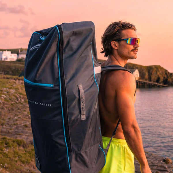 Man with sunglasses carrying a Bluefin SUP bag on his back by the water at sunset