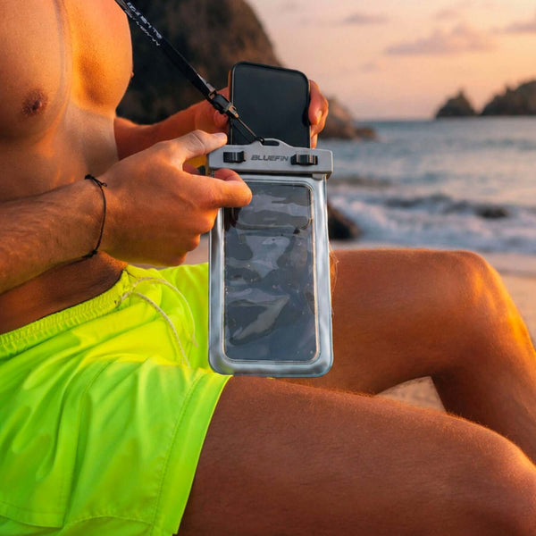 Person holding a waterproof phone case while sitting on a beach, wearing bright yellow shorts