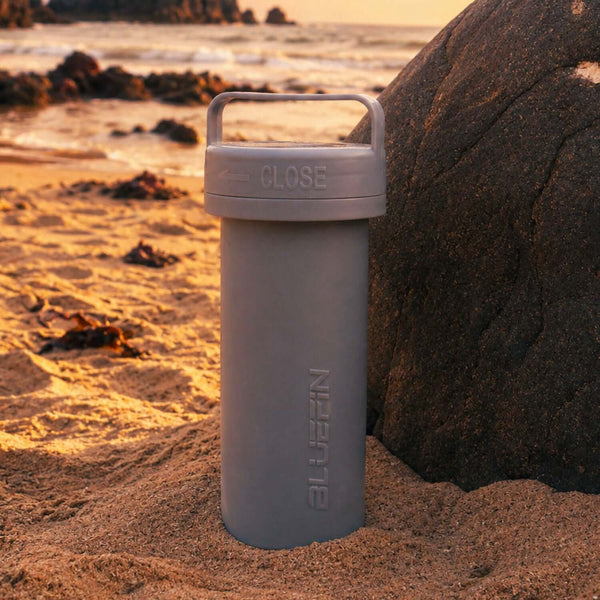 Gray waterproof container with a handle on sandy beach near a rock at sunset