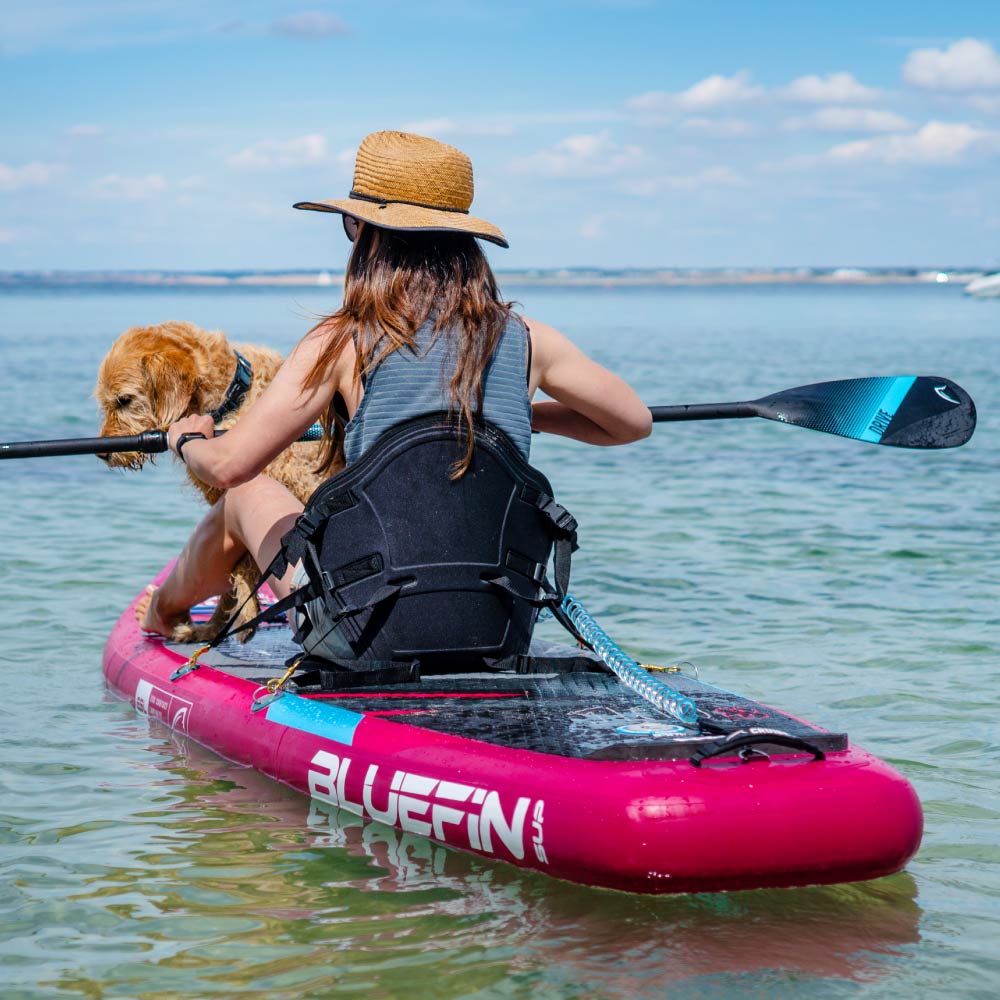 Person paddling a pink Bluefin SUP with a dog on a calm water surface under a clear sky.