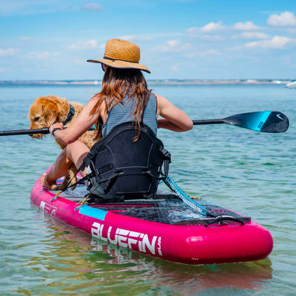 Person paddling a pink Bluefin SUP with a dog on a calm water surface under a clear sky.