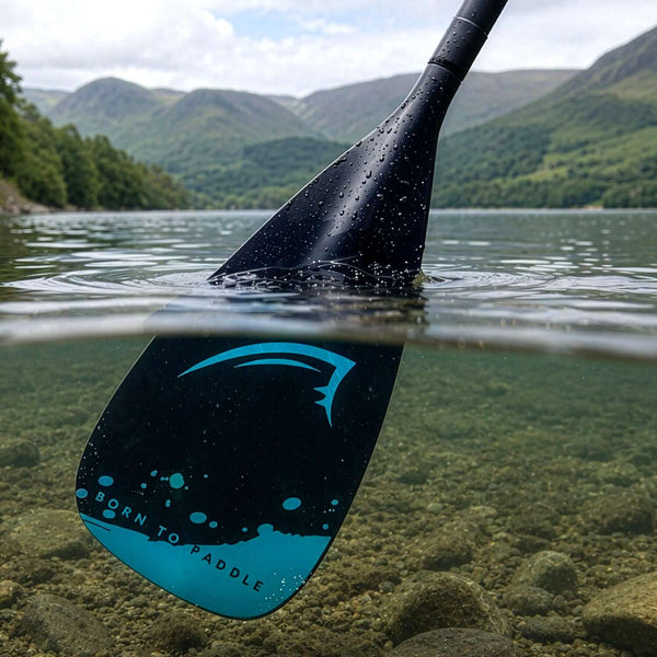 Close-up of a black paddle blade submerged in clear water, featuring blue design and text 'BORN TO PADDLE'.