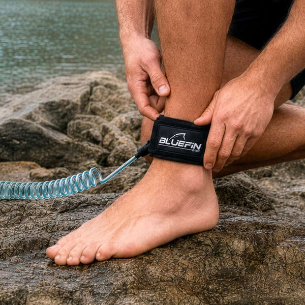 Person attaching a Bluefin SUP ankle leash to their ankle while standing on rocky shore