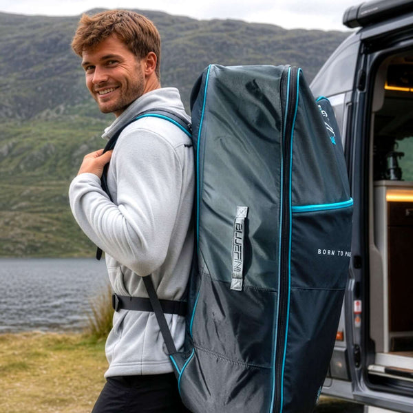 Man smiling while carrying a large dark backpack with blue accents near a lake and mountains