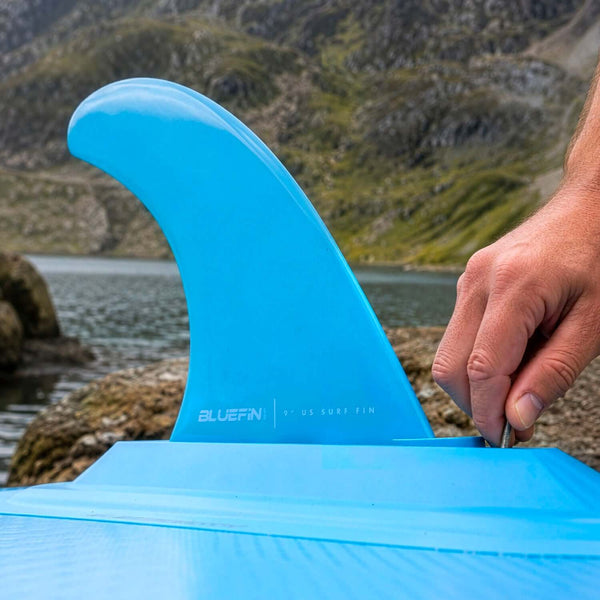 Bluefin 9" surf fin being attached to a blue paddleboard near a rocky shore and water.
