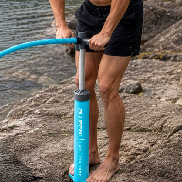 Person using a blue Bluefin Swift Pro Pump on rocky shore, barefoot, with water in background.