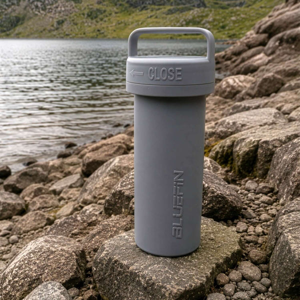 Gray cylindrical container with a handle, labeled 'CLOSE', on rocky lakeside.