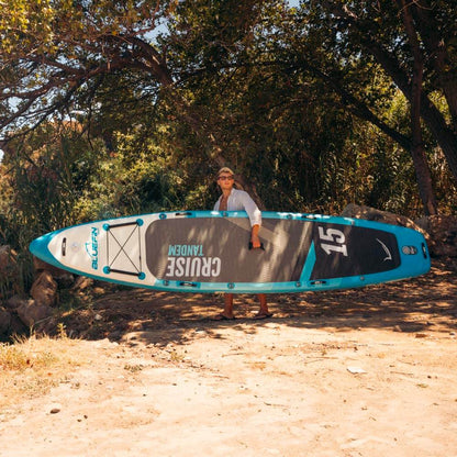 Person carrying a Bluefin SUP paddle board in a natural setting with trees and sandy ground.
