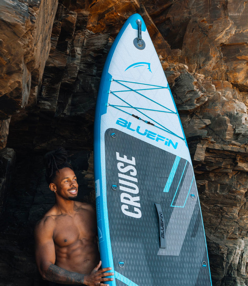 Inflatable Bluefin paddleboard with textured surface, held by a smiling man against rocky background