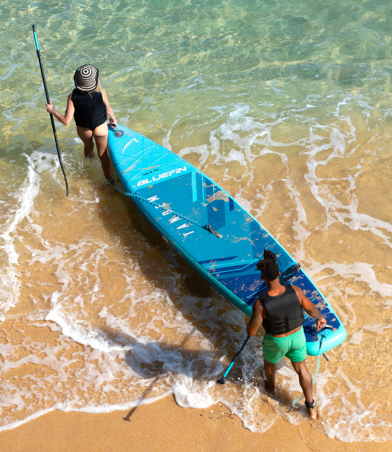 Two people walking into the water with a blue inflatable paddleboard and paddles, sandy beach background.