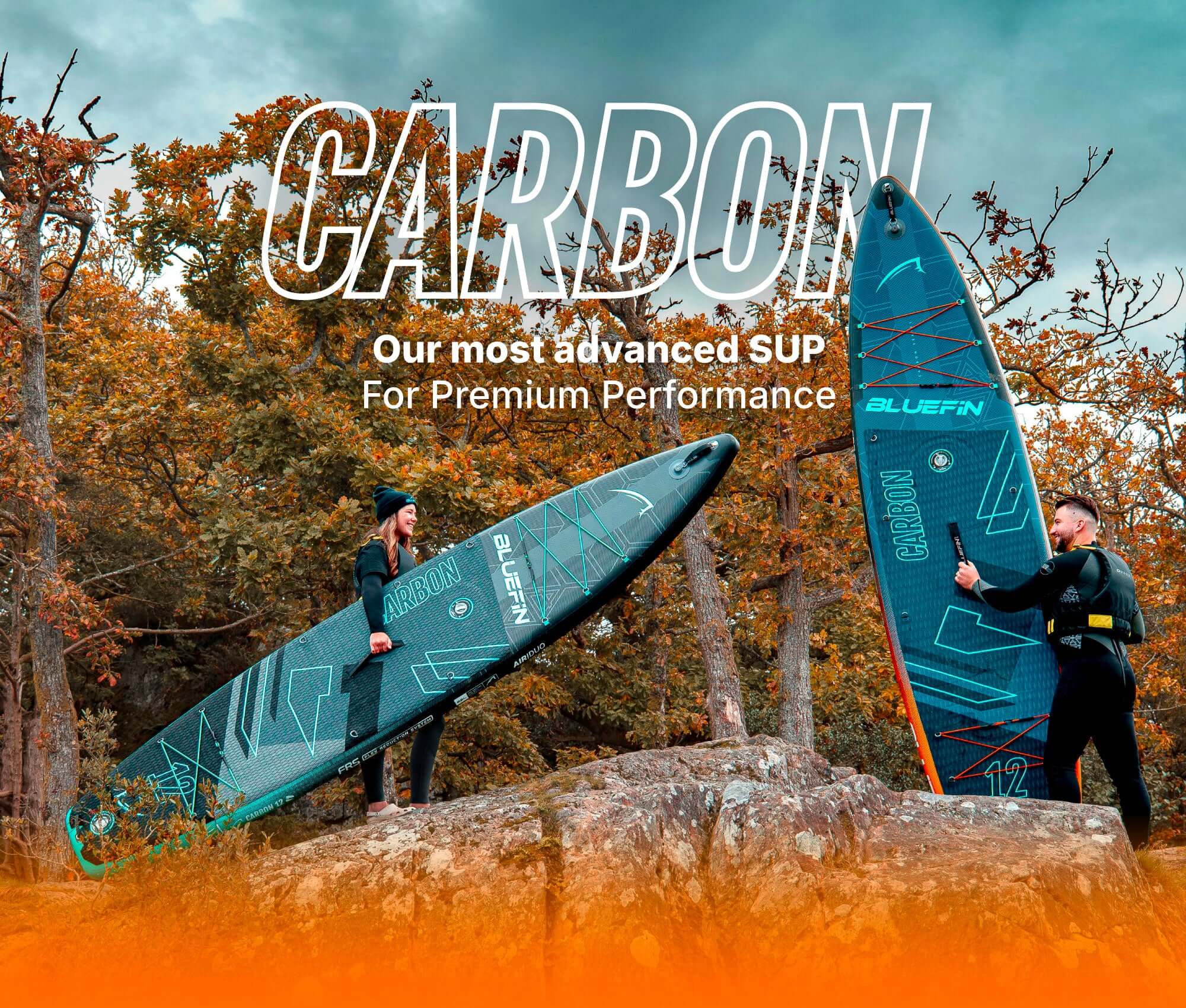 Two people holding inflatable paddleboards in a forest, one in black wetsuit, one in casual wear