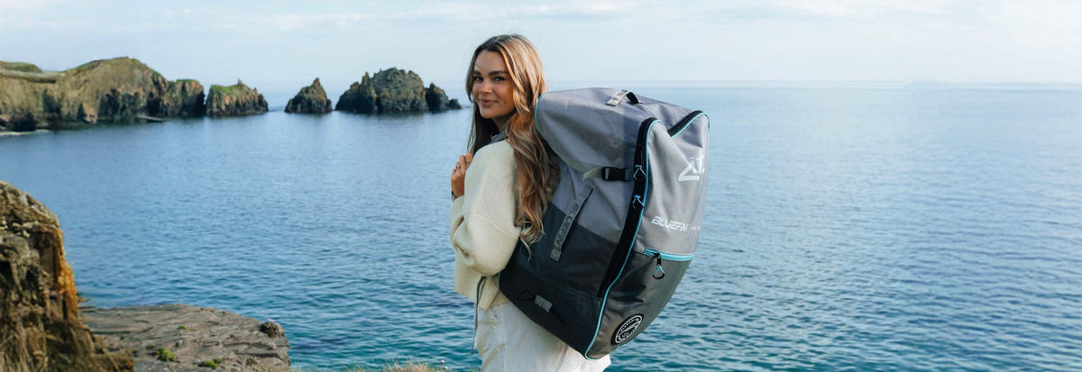 Woman wearing a cream sweater, carrying a large gray backpack by the water with rocky cliffs in the background.
