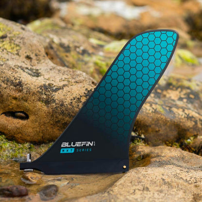 Bluefin NXT Series paddle board fin with teal honeycomb design on rocky surface.