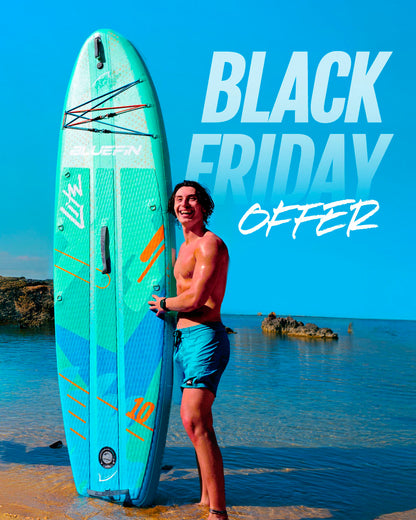 Person smiling while holding a blue-green inflatable paddleboard on a beach with clear water