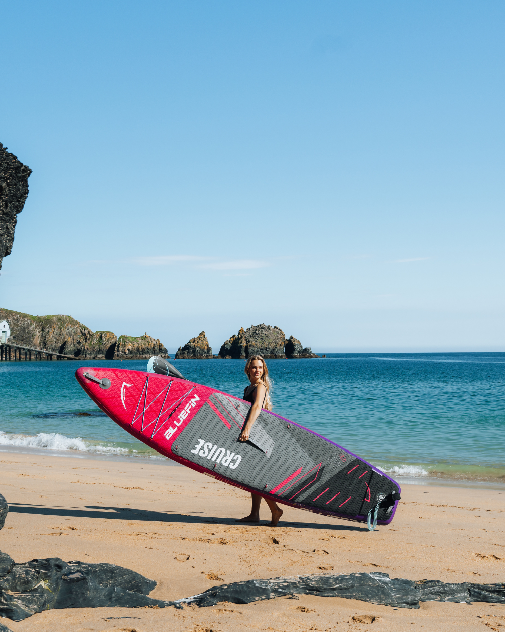 Woman holding a Bluefin inflatable paddleboard on a sandy beach with clear water and rocks in the background.