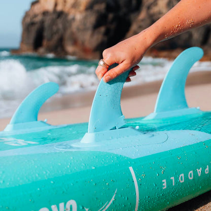Hand adjusting the fins on a turquoise inflatable paddleboard at the beach with waves in the background
