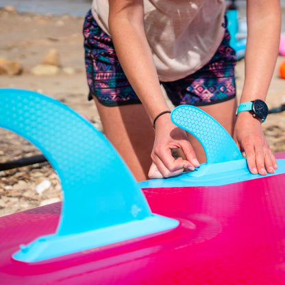 Person attaching a blue fin to a pink inflatable paddleboard on the beach