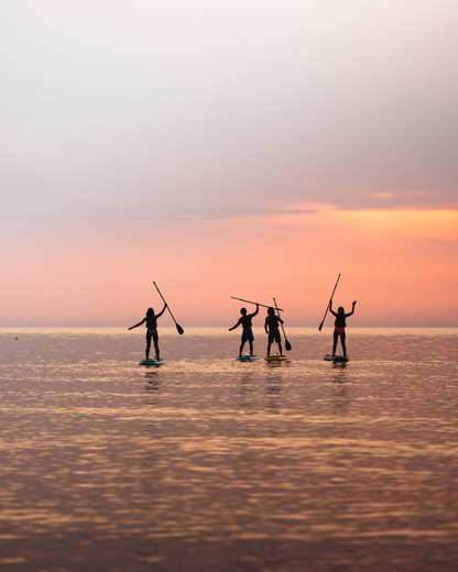 Four people paddleboarding at sunset, silhouetted against a colorful sky and calm water.