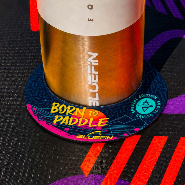 Colorful coaster with 'BORN TO PADDLE' text under a metal drink container on a textured surface