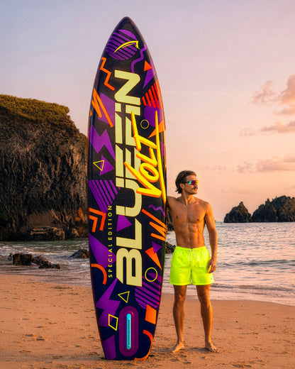 Colorful paddleboard with geometric designs, man in neon shorts standing on beach at sunset.
