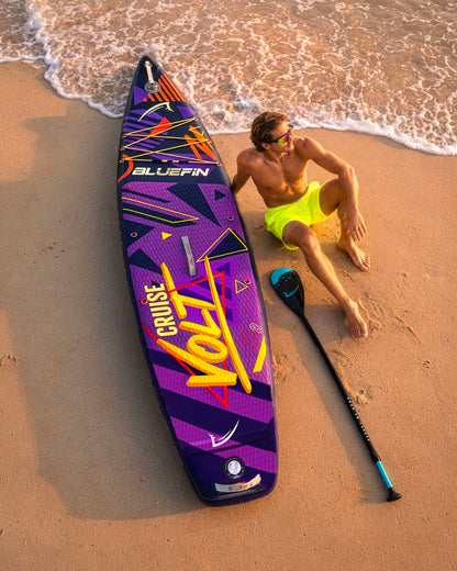 Inflatable paddleboard with colorful design, man in yellow shorts relaxing on sand, paddle nearby
