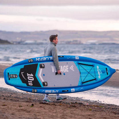 Person carrying a blue paddleboard on the beach with water in the background.