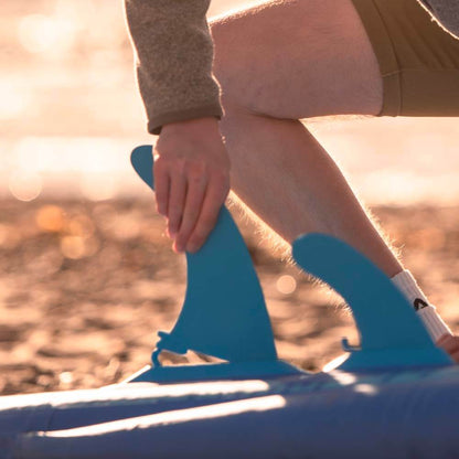 Person adjusting blue fins on a paddleboard by the shoreline during sunset.