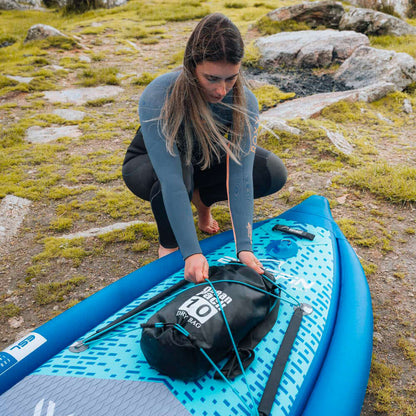 Woman securing a dry bag on a blue paddleboard in a natural outdoor setting.