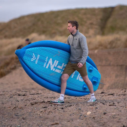 Person walking on the beach carrying a blue paddleboard, wearing shorts and a light jacket.